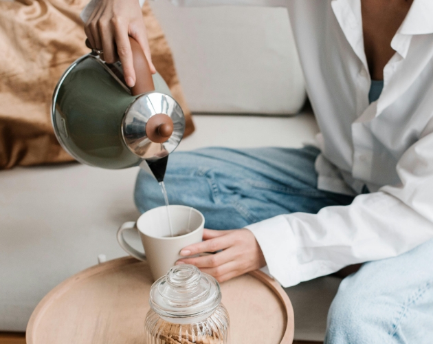 A woman sat on a sofa pouring hot water from an energy-efficient kettle into a mug placed on a
                   coffee table.